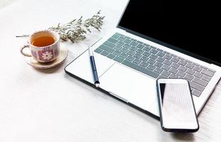 trabajo desde casa, oficina de trabajo en casa, computadora portátil o portátil con teléfono móvil con bolígrafo y taza de té china en el escritorio blanco de la oficina en casa. foto