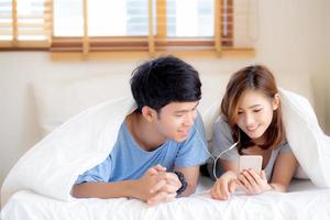 Young asian couple smile listen music with smart mobile phone on bed at bedroom with fun and enjoy, family sharing podcast or watching video from internet online with relax, lifestyle concept. photo