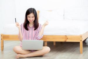 hermosa mujer asiática joven sentada usando tarjeta de crédito con computadora portátil, gesto de chica contenta ok y emocionada compra en línea con cuaderno en el dormitorio, concepto de estilo de vida. foto