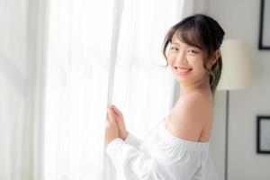 Portrait of beautiful young asian woman standing the window and smile while wake up with sunrise at morning, girl happy with freshness and cheerful, lifestyle and relax concept. photo