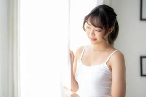 Portrait of beautiful young asian woman standing look at the window and smile while wake up with sunlight at morning, girl happy with fresh and cheerful, lifestyle and relax for wellness concept. photo