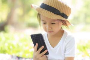 Beautiful asian child smiling using smart mobile phone in the garden, kid have passion playing game digital on smartphone on internet network in the park with happy, lifestyle and technology concept. photo