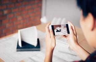 Architect man holding smartphone taking a photo of an architecture model sent to client and working in home office.