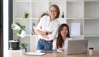 Two middle-aged women are looking at the camera while sitting on a desk in the office. female executives working in the office discussing documents photo