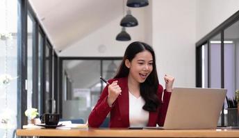 mujer feliz emocionada mirando la laptop celebrando una victoria en línea, joven asiática muy contenta gritando de alegría en la oficina foto