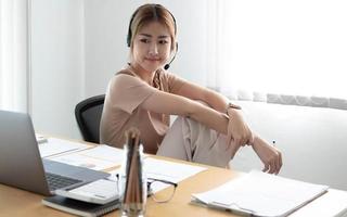 mujer de negocios asiática hablando con un equipo de colegas en una videoconferencia escribiendo una nota en el libro con cara de sonrisa. mujer que usa computadora portátil y auriculares para una reunión en línea. foto