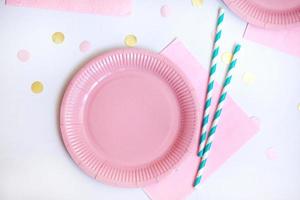 Pink paper disposable plate, napkin and straws for drinks on white background with confetti. Table setting for picnic. Bright eco-court. Birthday, party and holiday concept. Top view, flat lay photo