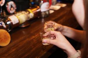 Two female hands clink glasses with alcoholic shots. Blurred bar background photo