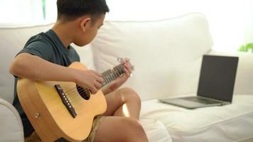 Asian boy learning to play the guitar in virtual meeting for play music online together with friend or teacher in video conference with laptop for online, Communication over Internet Learning concept