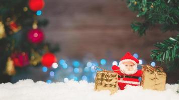 Santa Claus doll with a gift box placed on the snow and the Christmas tree background with bokeh and wood.selective focus photo