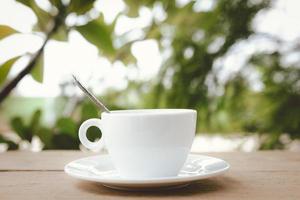 A white coffee cup with spoon in a cup and saucer are placed on a wooden plate and on the landscape nature background. photo