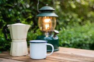 enfoque selectivo taza de café de esmalte blanco y juego de café en el jardín con farolillos antiguos en un ambiente de camping. enfoque suave. foto