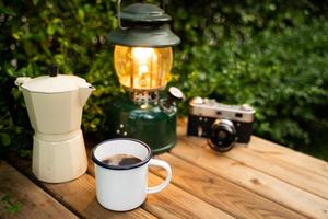 enfoque selectivo taza de café de esmalte blanco y juego de café en el jardín con farolillos antiguos en un ambiente de camping. enfoque suave. foto