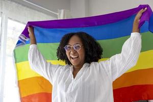 niña afroamericana sosteniendo la bandera del arco iris lgbtq en su dormitorio por salir del armario y la celebración del mes del orgullo para promover la diversidad sexual y la igualdad en el concepto de orientación homosexual foto