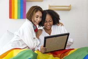 Couple of same sex marriage from difference races with LGBTQ rainbow flag looking at their photo during pride month to promote equality and differences of homosexual and discrimination concept