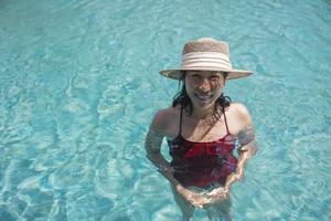 feliz mujer asiática en traje de baño rojo y un sombrero de paja relajándose en la piscina mirando a la cámara junto a la piscina en koh mak, phangnga, tailandia. relajarse y viajar concepto. confort resort vacaciones de verano foto