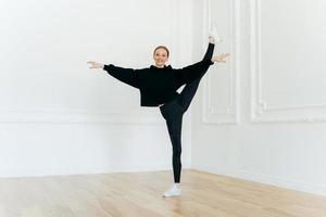 Satisfied female ballerina balances on one leg, stretches in studio, practices yoga, demonstrates good flexibility, dressed in black clothes, has happy expression, performes exercise indoor. photo