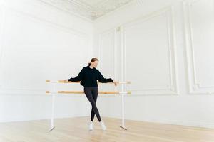 el bailarín de ballet clásico tiene ejercicios de estiramiento, se para cerca de las barras de ballet, usa sudadera negra y polainas, hace ejercicios en el estudio, se enfoca hacia abajo. instructora certificada de coreografía foto