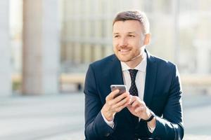 Smiling male manager with appealing look, dressed formally, chats with costumer, uses modern smart phone, sits against urban background, connected to wireless high speed internet. Technology photo