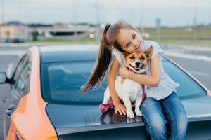 Photo of merry small female kid has pony tail embraces with love her favourite pet, pose at trunk of automobile, play together, walk in open air, enjoy togetherness. Children and animals concept