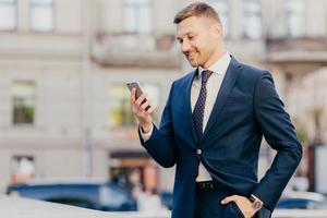 Horizontal shot of happy businessman keeps hand in pocket, wears formal suit and wristwatch, recieves notification on smart phone, finds out about high financial balance. Cheerful trader outdoor photo