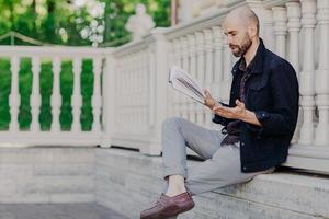 Attractive bald man reads attentively interesting book, gestures with hand, has stunning plot, tries understand feeling of main characters, poses outdoor, enjoys fresh air and wonderful view photo