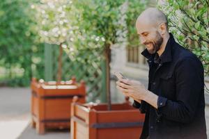 Horizontal shot of happy bald male hipster with beard, dressed in black jacket, holds smart phone, reads information or news on web page, enjoys free internet, strolls outdoor in beautiful park photo