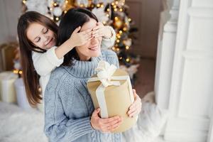 una pequeña y adorable niña prepara una sorpresa para su madre, cierra los ojos y le da un regalo envuelto como soporte en el fondo de año nuevo. bonita mujer recibe un regalo de su hija. concepto de sorpresa foto