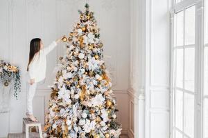 Horizontal portrait of small female child stands on chair, decorates Christmas tree, tries to show best, being at home, enjoys domestic calm atmosphere, wants to satisfy parents, being hard working photo