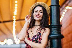 Excited female with dark hair, shinig eyes and red lips, dressed in dress and summer hat, posing at camera with happy and confident expression. Attractive female brunette with make-up resting at cafe photo