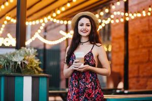 Positive woman with dark hair and appealing face, wearing summer dress and hat, holding takeaway coffee while standing at outdoor cafe, having happy expression. Beautiful woman resting at cafe photo