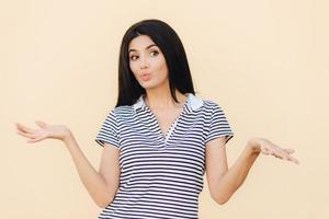 Beautiful female has clueless expression, shrugs shoulders, keeps lips round, looks hesitant aside, dressed in casual clothes, isolated over studio background. People and uncertainty concept photo