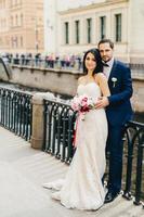 Handsome bearded bridegroom embraces his bride in white dress with bouquet stand together at bridge, have good mood after registration of their marriage. Good relationships concept photo