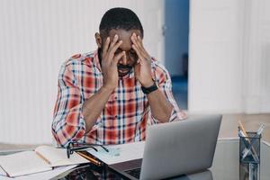 el hombre afroamericano agotado sufre dolor de cabeza en la computadora portátil. estrés en el trabajo, desgaste laboral foto