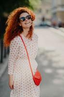 Optimistic foxy woman with glad expression, looks into distance, wears shades, long dress, strolls outdoor on street, enjoys nice summer day, has broad smile. People, lifestyle, rest and lifestyle photo