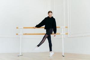 Young ballet dancer has warm up, stands near handrails, dressed in black sportclothes, stands on tip toe, wears white sneakers, focused down on floor, has cheerful facial expression, raises leg photo