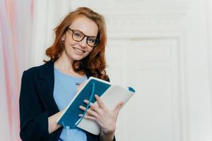 Prosperous satisfied businesswoman in elegant clothes writes in diary, has glad expression, wears spectacles, makes list of plans, writes text message in notebook, stands indoor. Female makes notes photo