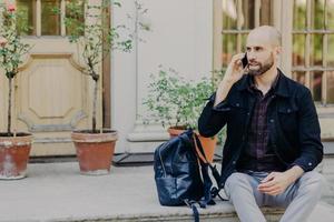 Bearded male tourist with thick beard, has phone conversation, has rest after excursion, uses modern cell phone for communication, looks pensively aside, admires wonderful scenery. Technology concept photo