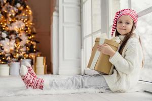 Portrait of adorable pleasant looking girl wears comfortable warm clothes, holds wrapped gift box, recieves congratulations with New Year from parents and relatives, has pleased expression photo
