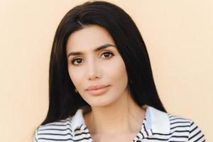 Close up portrait of brunette young female with healthy clean skin, perfect make up, demonstrates her natural beauty, looks directly into camera, dressed in striped dress, isolated on light background photo