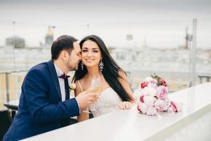 Romantic newlyweds drink champagne, clink glasses, being very happy to celebrate their wedding, feels passion and great love to each other. Beautiful bride recieves kiss from bridegroom