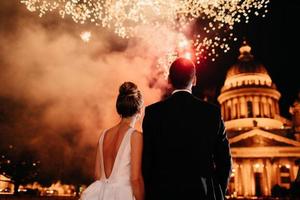 Back view of lovely married couple stand backs to camera, admire beautiful firework in night sky. Bride and groom stand closely to each other, watch salute, celebrate their wedding. Nice moment photo