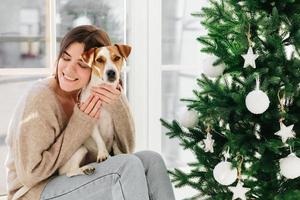 toma horizontal de una mujer sonriente cariñosa abraza al cachorro con amor, disfruta de un ambiente acogedor y un momento encantador, sonríe agradablemente, decora el árbol de año nuevo juntos, celebra la fiesta favorita en casa foto