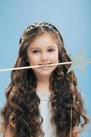 Photo of attractive woman kid with pleasant appearance, has long dark hair, holds magic wand in mouth, poses over blue background, dressed in princess dress. Vertical image. Childhood concept