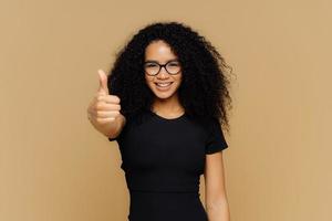 Waist up shot of satisfied supportive woman shows thumb up, cheers best friend, encourages for excellent efforts, wears glasses and black t shirt, stands against beige background. Body language photo