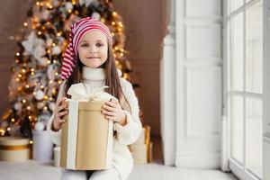 Indoor shot of pleasant looking small kid with blue charming eyes, wears santa hat, holds present in wrapped box, sits over decorated New Year background. Childhood, celebration concept photo