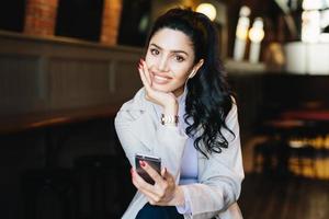 Caucasian young brunette woman with long pony tail having pretty face and gentle hands with red manicure listening to her favorite music with earphones and cell phone posing at cafe background photo