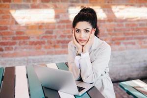 dama atractiva con cabello oscuro y maquillaje, largas uñas rojas sosteniendo sus manos en las mejillas descansando en un café sentado en la mesa frente a una laptop abierta esperando una videollamada de sus familiares o amigos foto