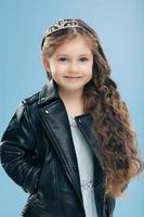 Vertical shot of attractive small female child has long curly hair, wears crown and leather jacket smiles positively isolated over blue wall. Happy little girl dressed in stylish clothes models indoor photo