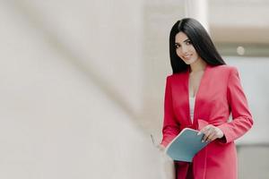 Beautiful smiling lady in elegant red costume, writes some notes in notepad, stands outside, copy space on left side for your promotional content. Businesswoman has meeting with partners or colleagues photo